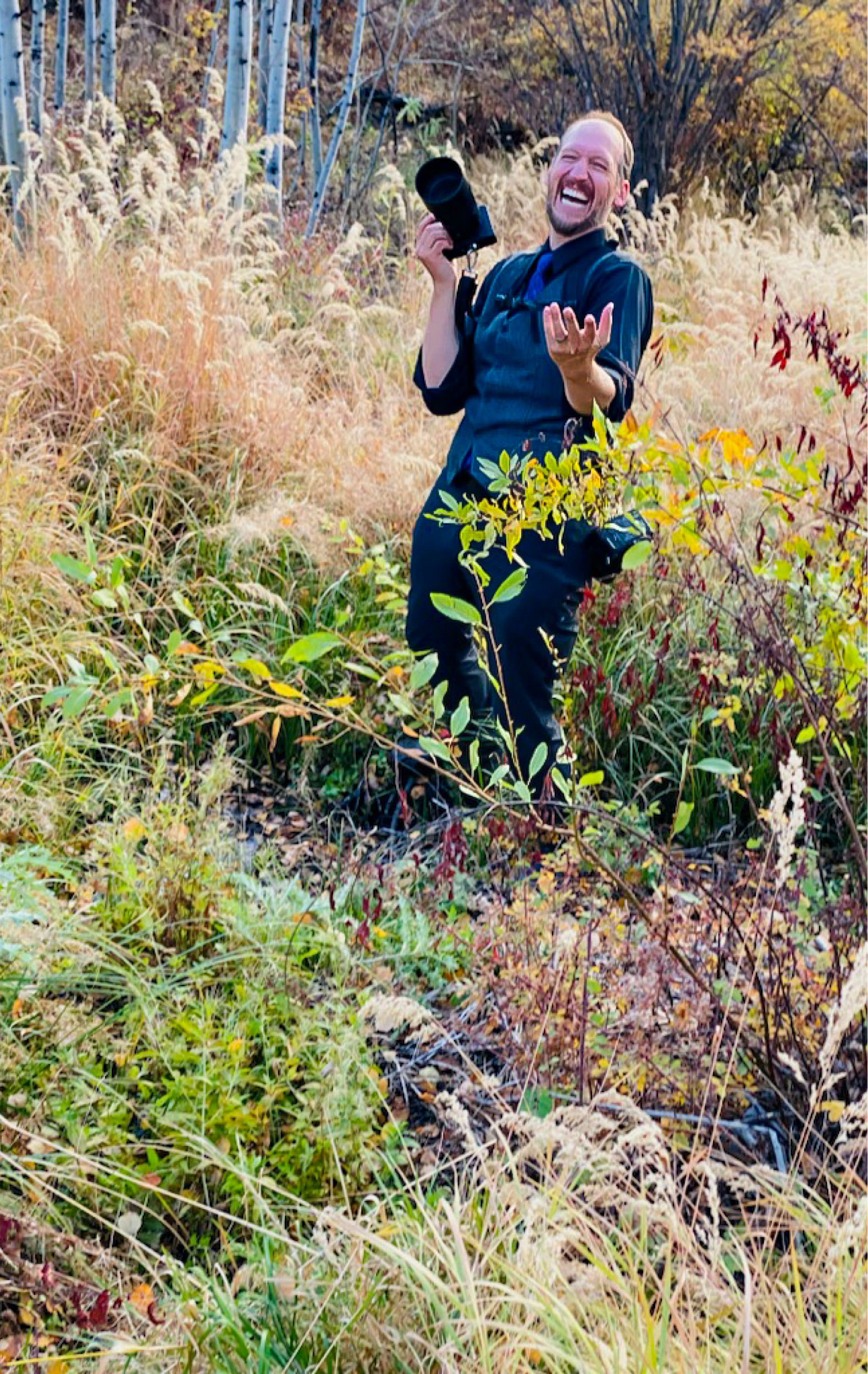 Jeff laughing with his camera raised in a field of autumn grasses